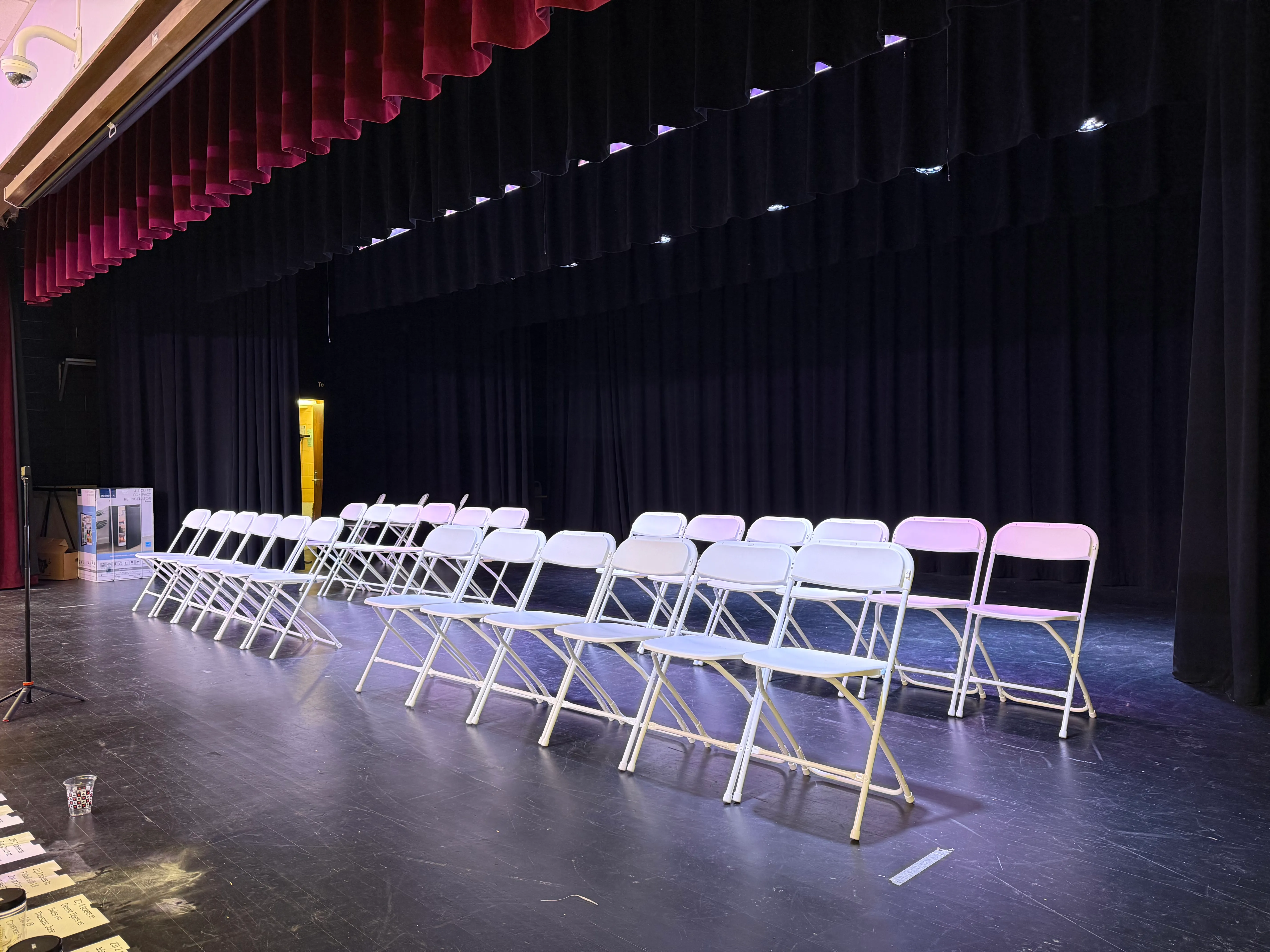 An empty stage before doors: two rows of chairs set and lit, set list taped to the floor